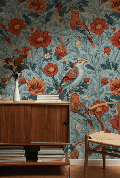 A wooden sideboard and woven chair sit in front of Sienna Songbird Wallpaper, which features blue florals, red birds, and botanical blooms. A white vase with dried leaves and stacked magazines complete the nature-inspired decor.