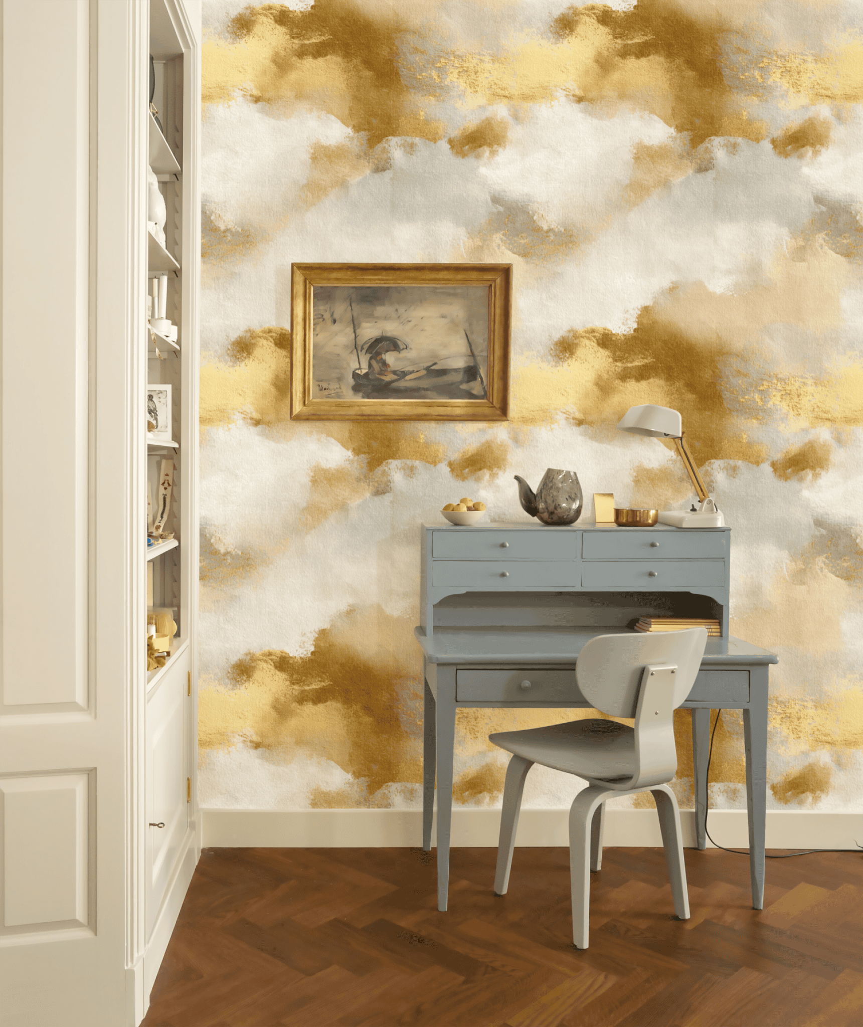 A small blue writing desk with a chair is set against Golden Haze Wallpaper. A painting, lamp, and decorative items rest on the desk, while bookshelves are visible to the left.
