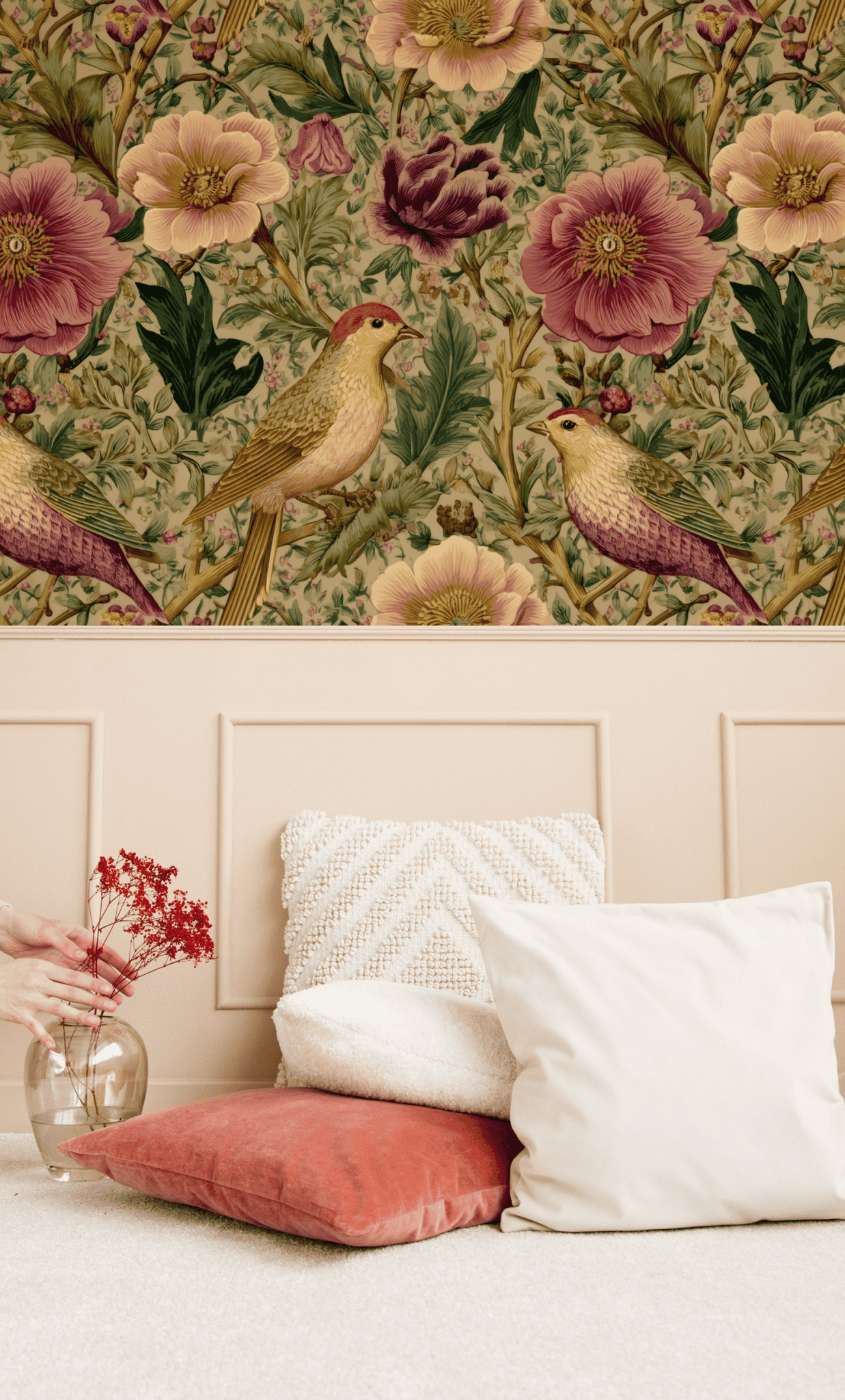 The Birdsong Bloom Wallpaper above a beige panel wall, paired with white and coral cushions and a hand holding a vase with red flowers, creates a tranquil indoor garden scene.