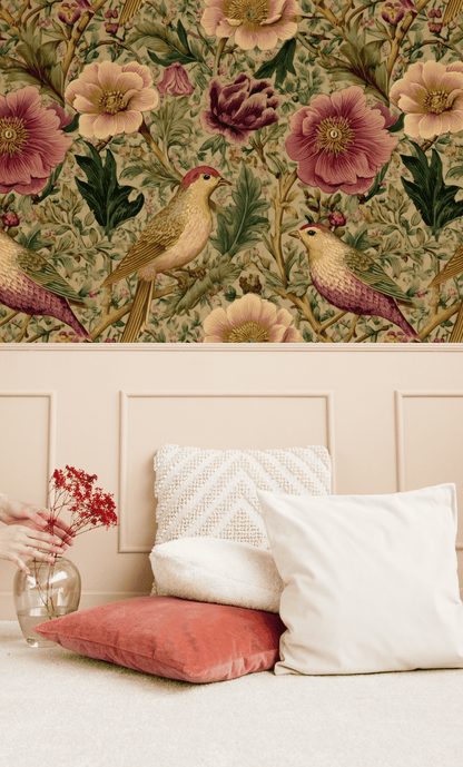The Birdsong Bloom Wallpaper above a beige panel wall, paired with white and coral cushions and a hand holding a vase with red flowers, creates a tranquil indoor garden scene.