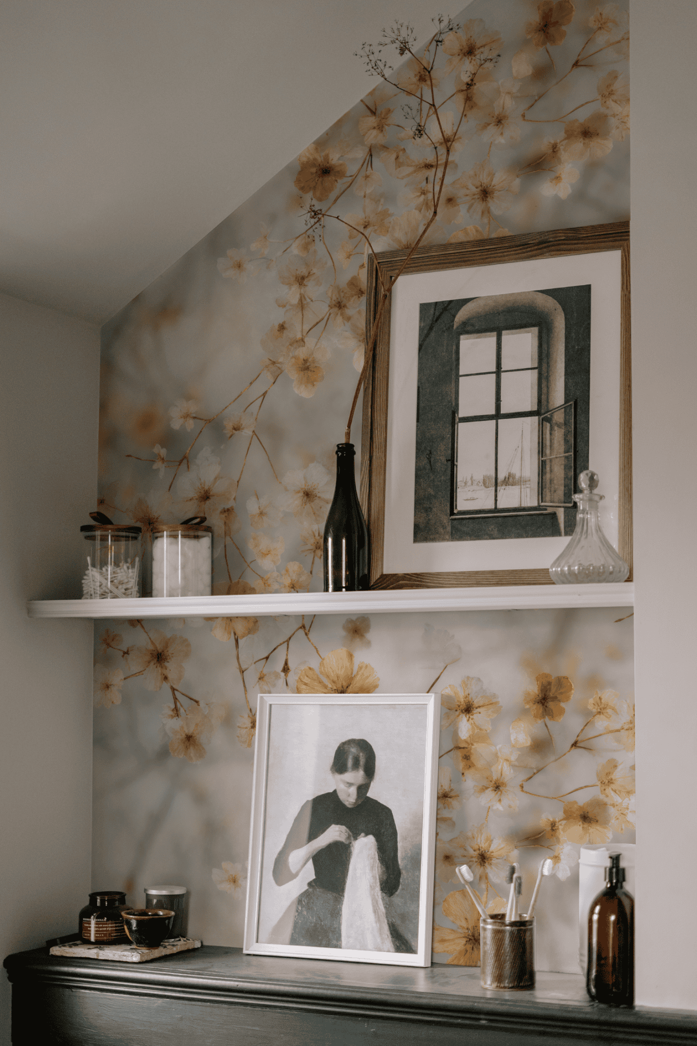 A shelf with framed art, bottles, jars, and brushes is displayed against a wall adorned with Whispering Petals Wallpaper, adding tranquil decor to the space.