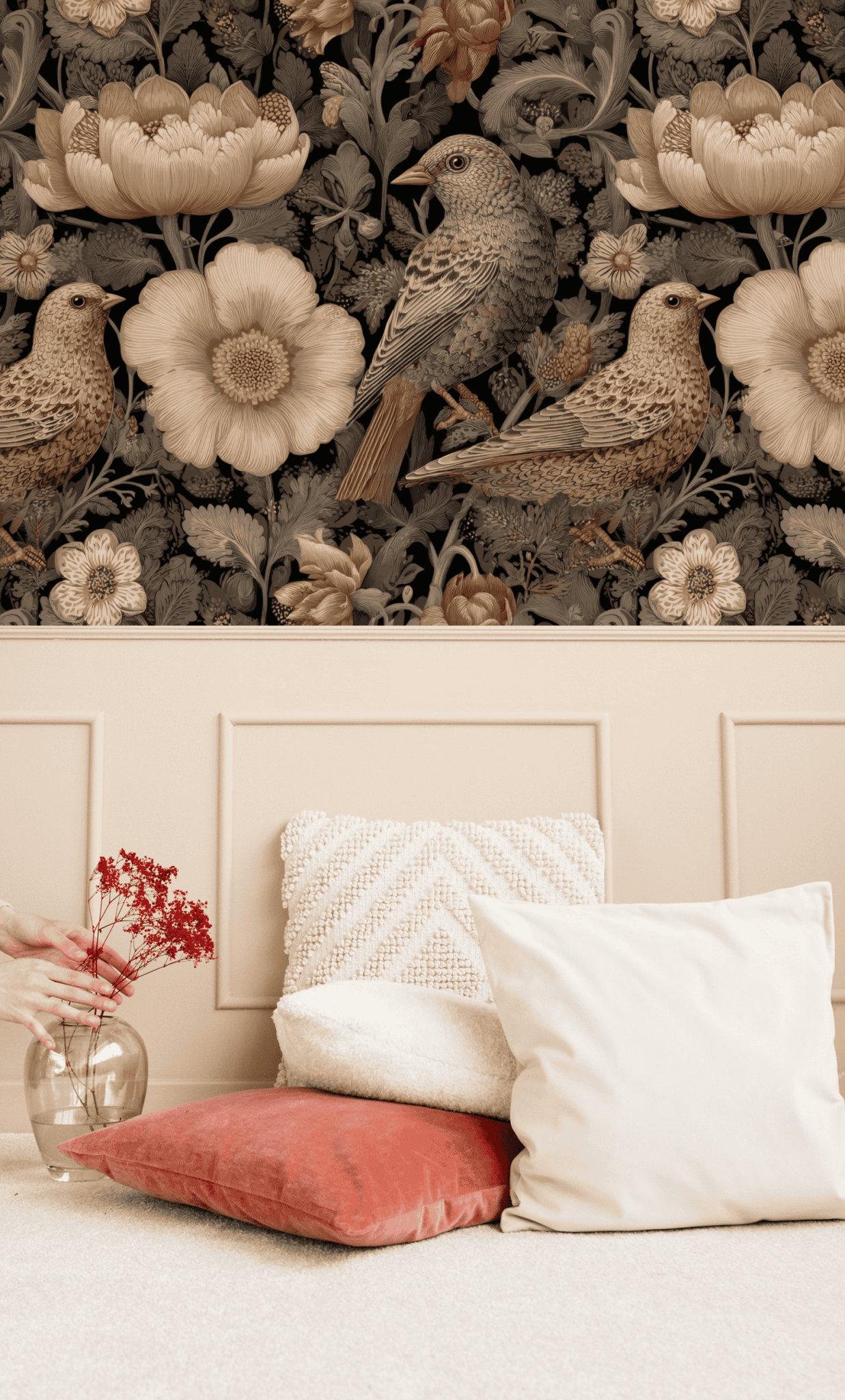 A hand places a glass vase with red dried flowers by white and red cushions; Auburn Songbird Wallpaper with graceful birds and muted floral patterns decorates the background.