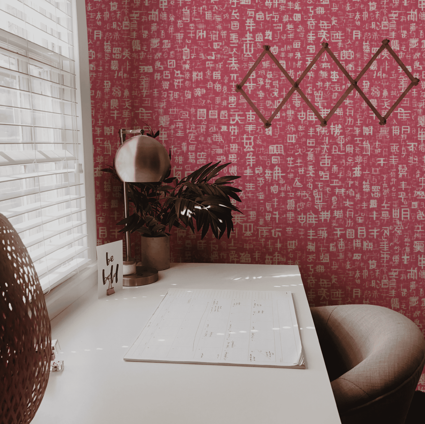 A modern desk setup with a plant, lamp, notepad, and "be still" card against a pink backdrop featuring Script Harmony Wallpaper and geometric wall decor.