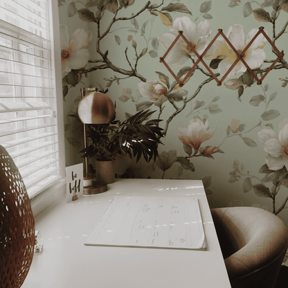 A white desk with a planner, lamp, plant, and "be still" card sits by a window; Magnolia Blossoms Wallpaper and a folded wooden rack add refined tranquility to the wall behind.
