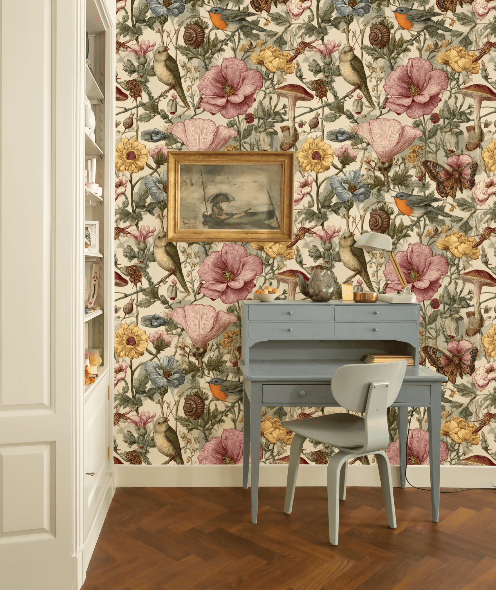 A small blue desk and chair sit against a wall adorned with Verdant Whimsy Wallpaper, featuring birds and blooms, while framed art and decor items are displayed above and on the desk.
