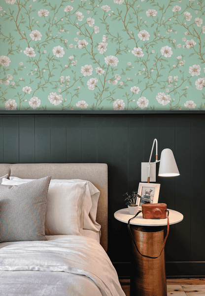 A neatly made bed with neutral bedding sits beside a round white nightstand topped with a lamp, books, and decor, set against Whispering Blooms Wallpaper featuring white flowers above rich dark green wainscoting.