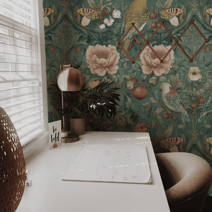 A white desk with a lamp, chair, notepad, and “be still” card sits by a window; the wall features vibrant birds on Verde Aviary Wallpaper with geometric mirrors—a true botanical escape.