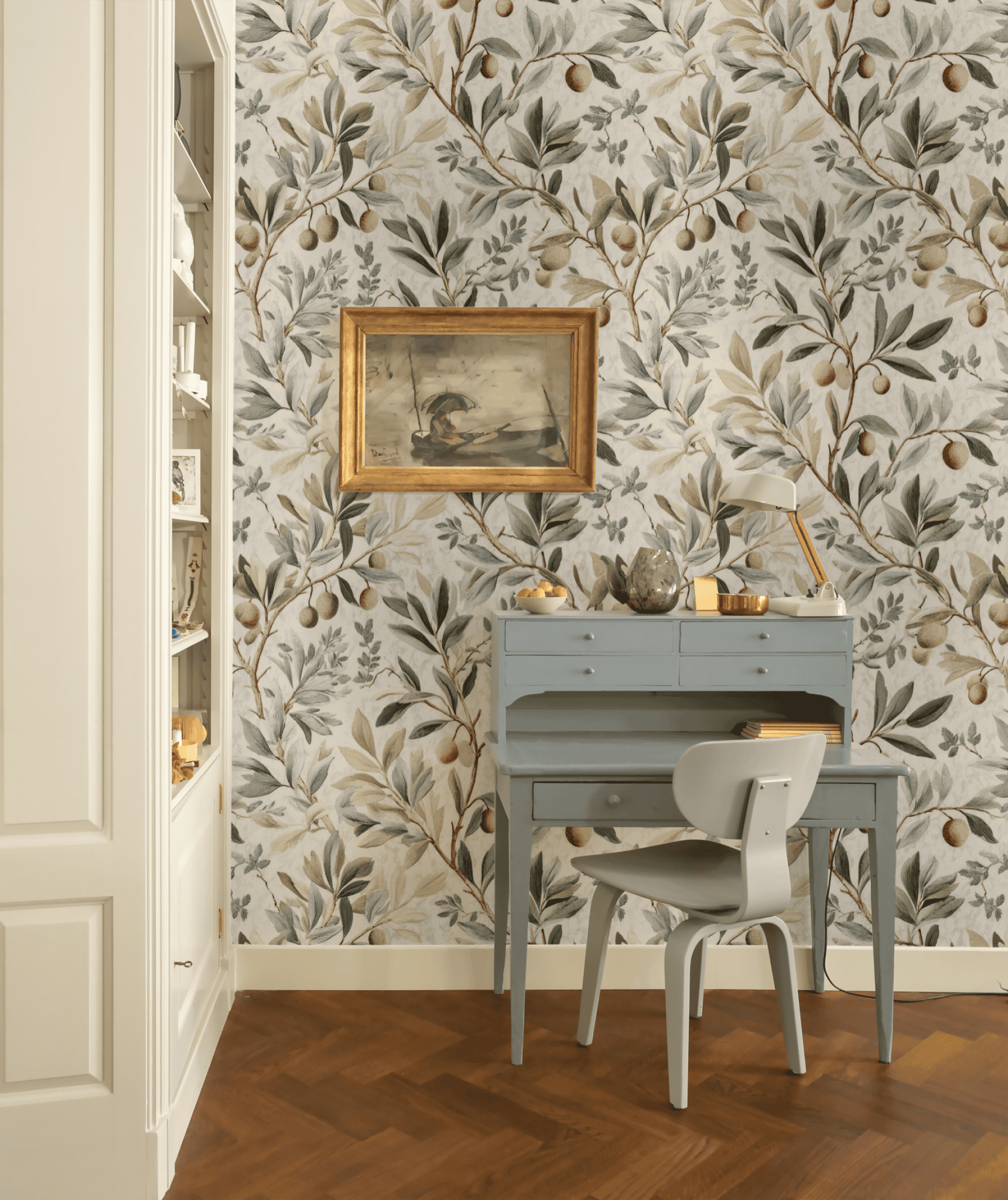 A small blue desk with a matching chair sits against Olive Grove Wallpaper; a framed painting hangs above and a desk lamp rests on the desk.