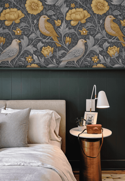 Create a sophisticated bedroom ambiance with the Golden Aviary Wallpaper, featuring golden and grey birds and blossoms on a dark background, paired with a bed, striped and white pillows, and a round side table with lamp and camera.