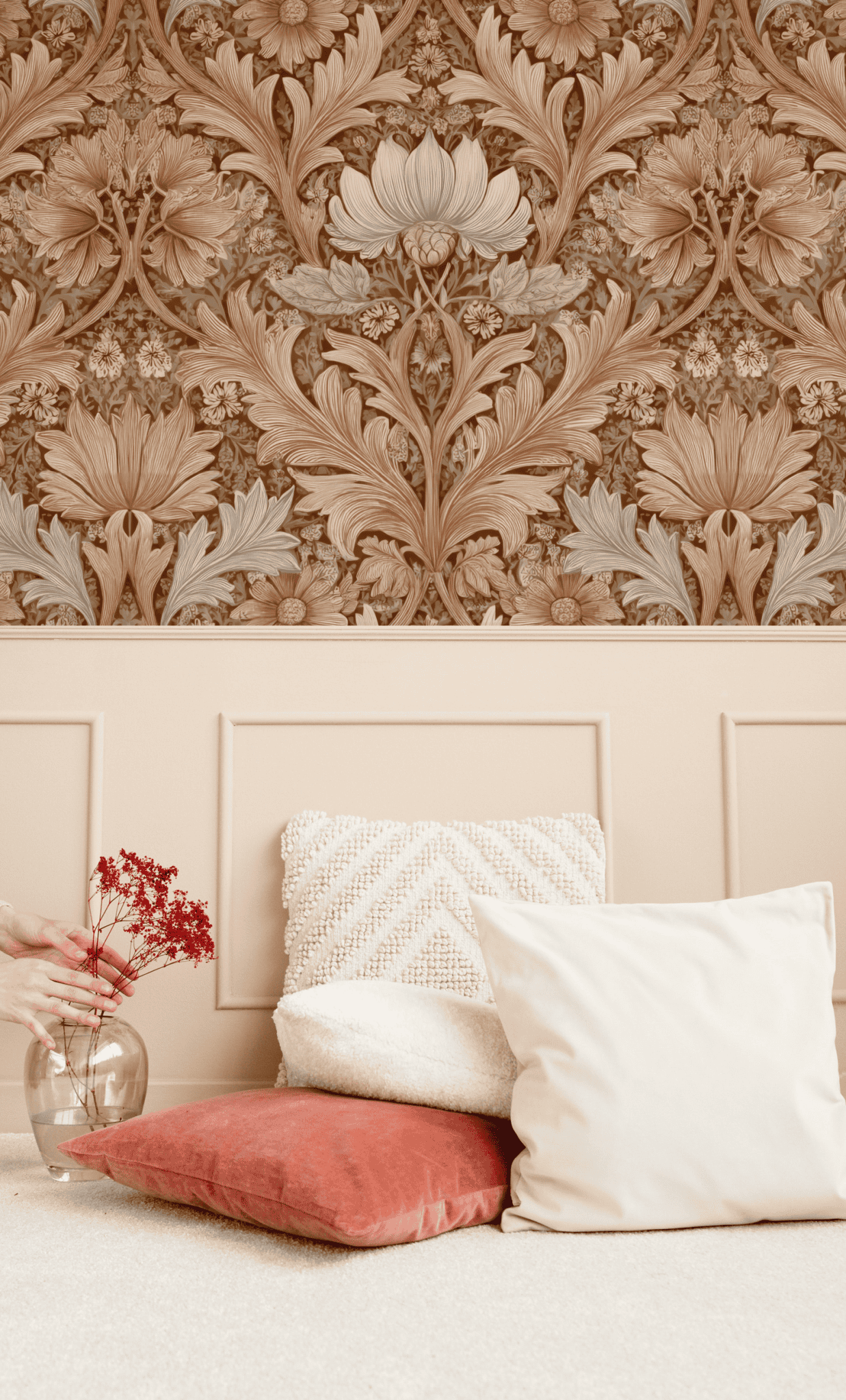 Antique Flora Wallpaper above beige wainscoting exudes timeless charm, while white and red pillows and a hand arranging red flowers in a glass vase complete the elegant scene.