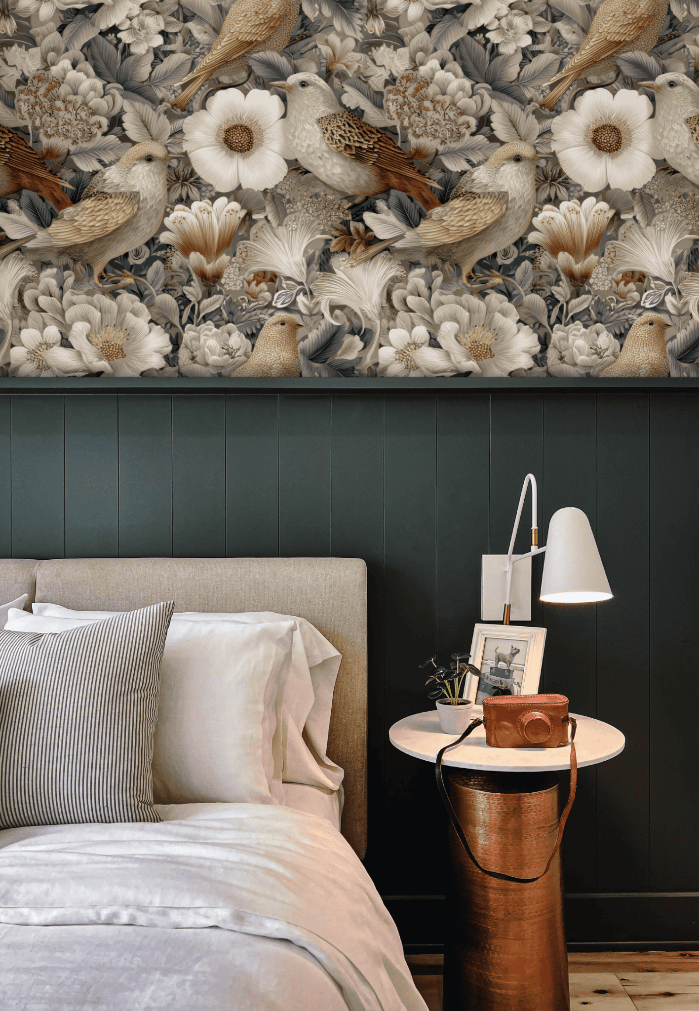 A bedroom featuring a beige upholstered bed, striped pillows, a round side table, and an accent wall with Aviary Harmony Wallpaper, adorned with delicate birds and flowers for a serene, nature-inspired look.